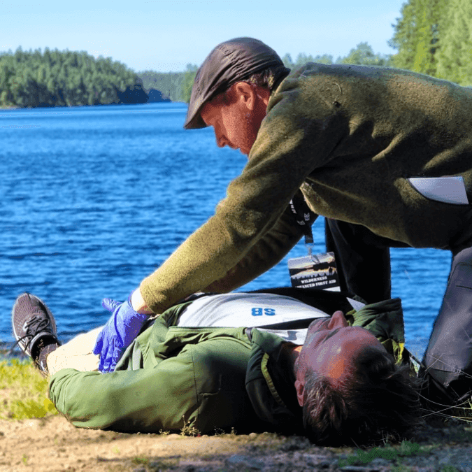 Man i gröna kläder undersöker en annan man som ligger på en strand med sjö i bakgrunden.