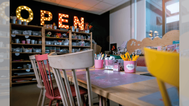 En skylt med lampor som bildar ordet open. Stolar vid ett bord, i bakgrunden en hylla med material för att skapa.