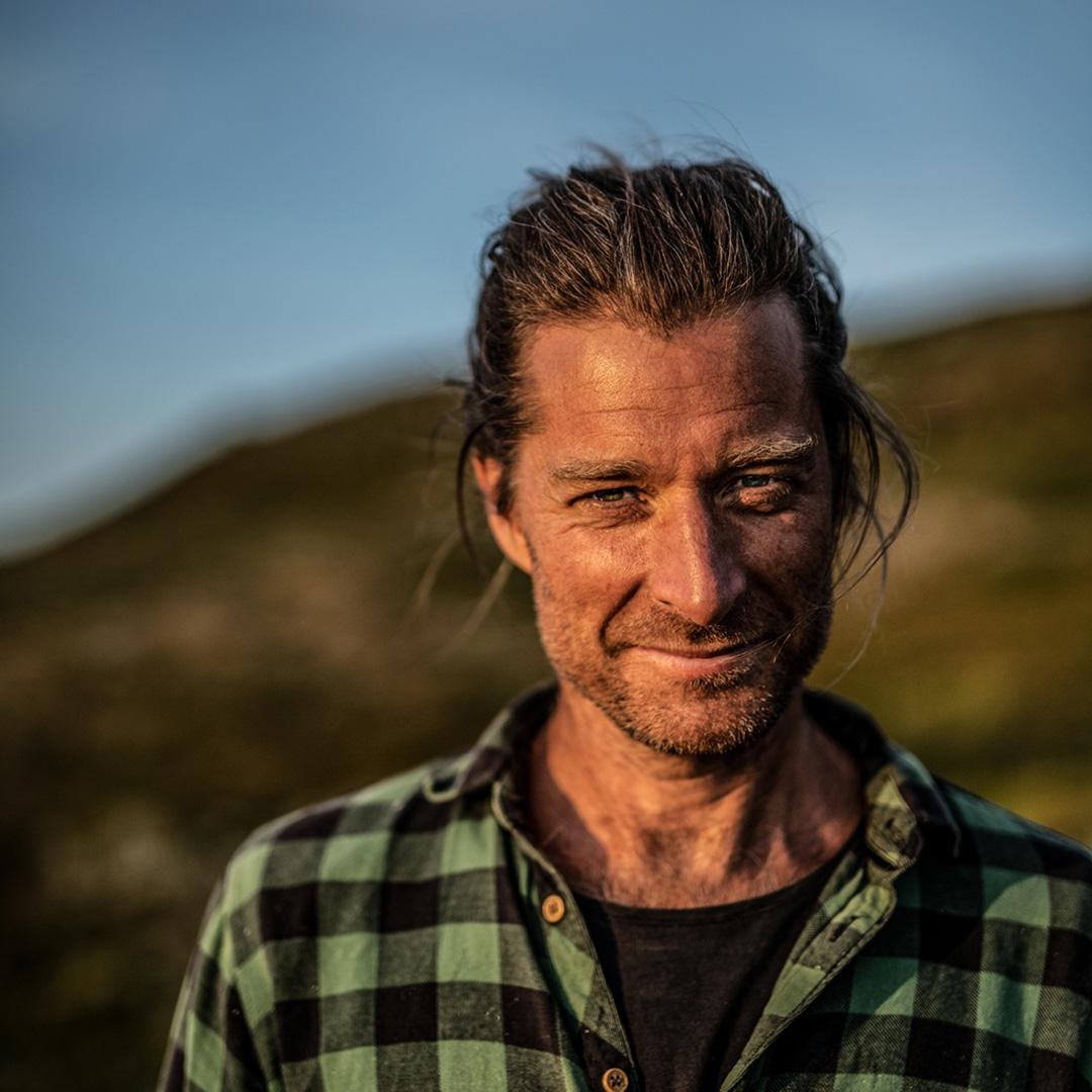 Porträtt av Markus Torgeby utomhus i naturligt ljus, med uppsatt hår och skäggstubb, iklädd grönrutig skjorta. Bakgrunden är ett öppet landskap med himmel och natur.
