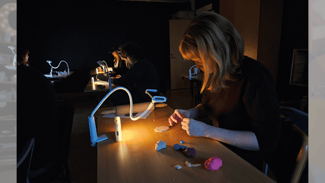Flicka sitter i ett mörkt rum med en kamera vid bordet och pysslar - troligtvis skapar hon stop-motionfilm.