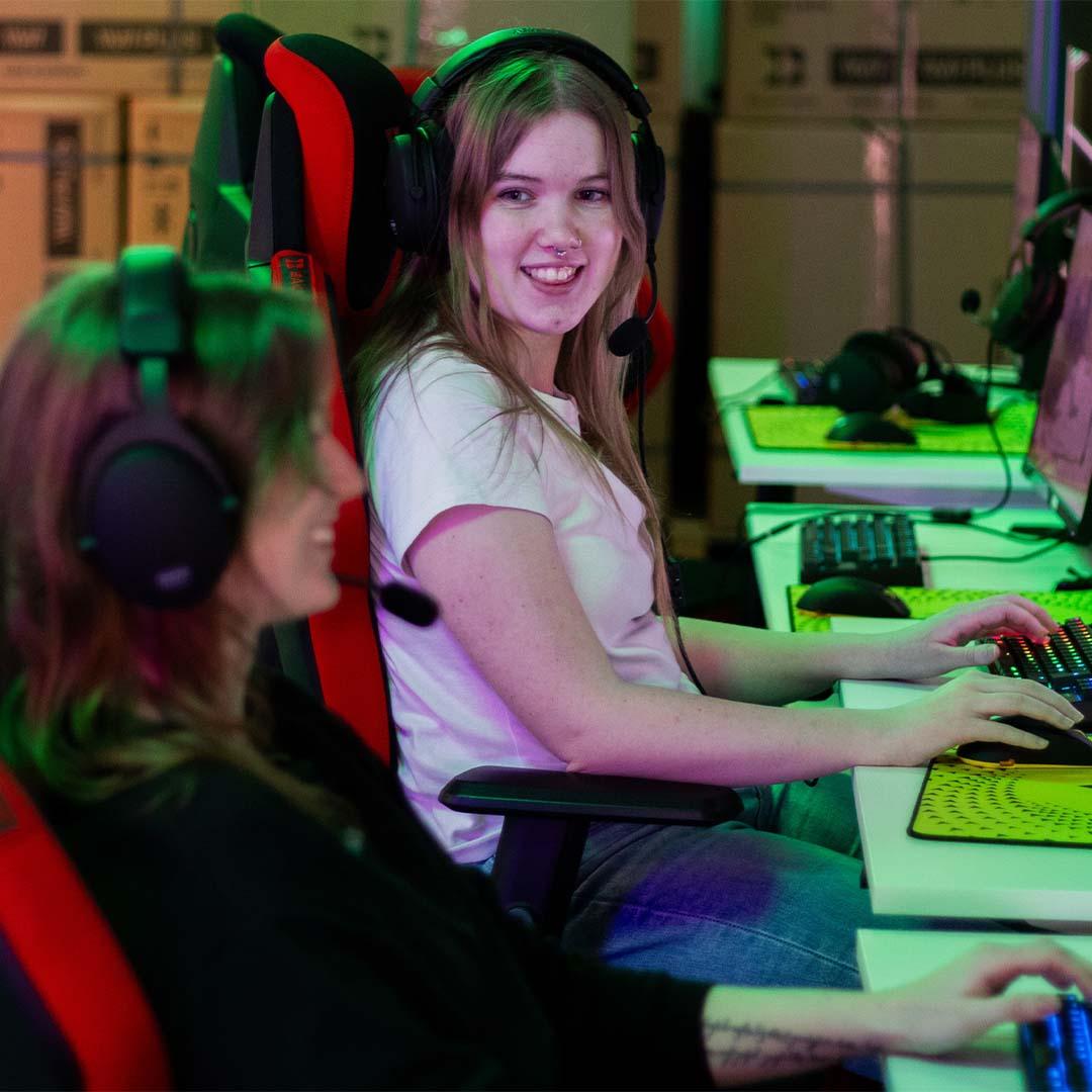 Två tjejer med hörlurar sitter vid datorer och gamear