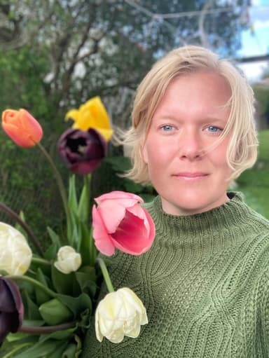 En tjej står vid blommor och tittar rakt in i kameran.