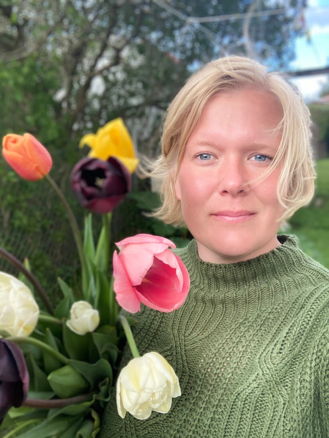 En tjej står vid blommor och tittar rakt in i kameran.