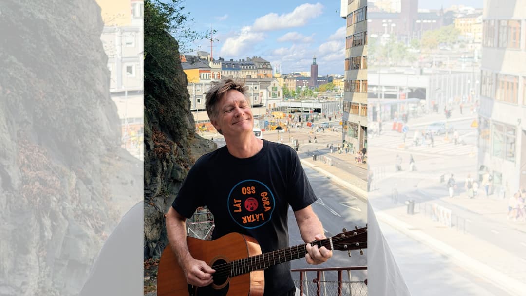 En man håller i en gitarr i stadsmiljö med ett stort leende på läpparna. På hans T-shirt står det "Låt oss göra låtar" i en cirkel.