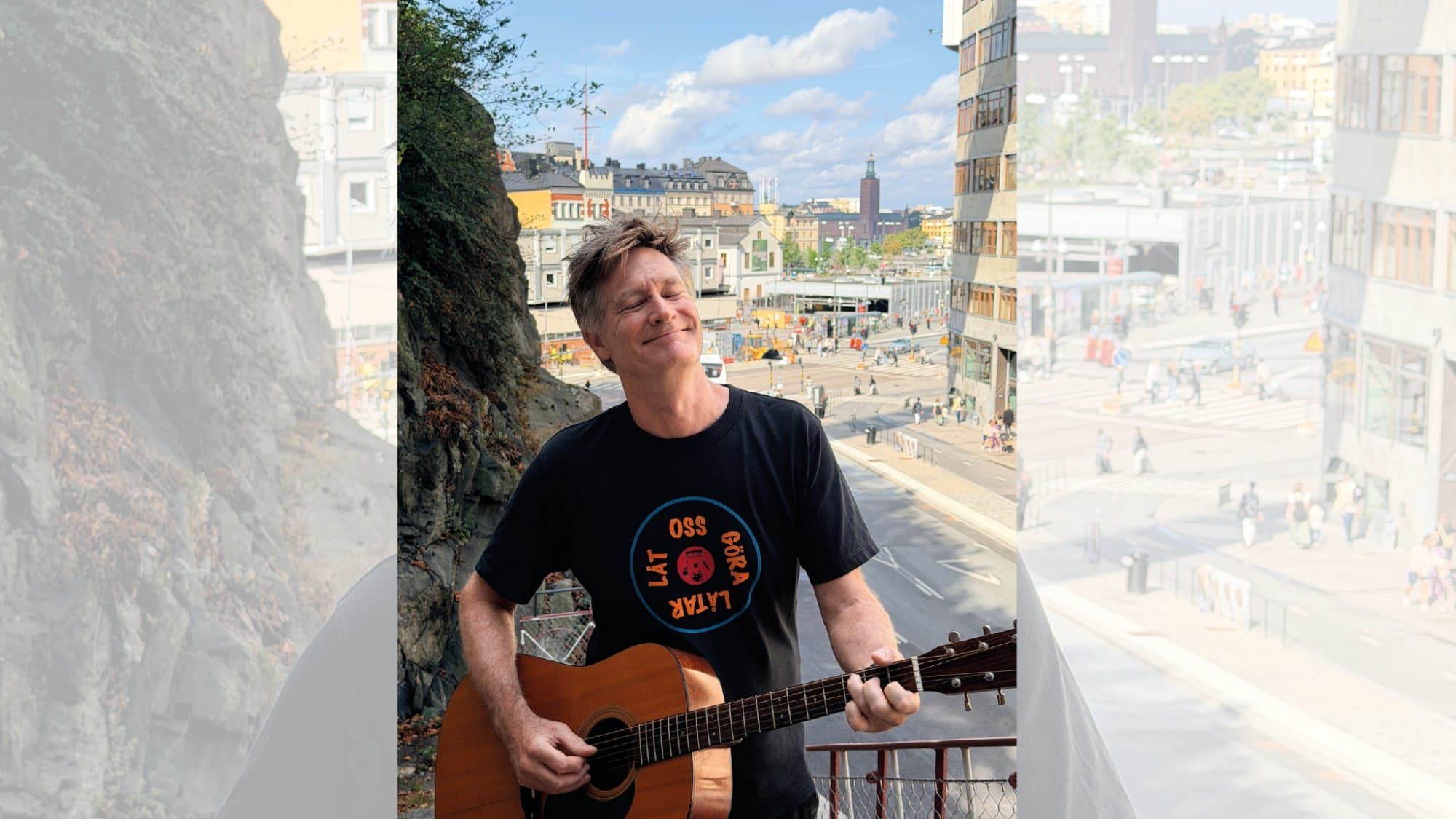 En man håller i en gitarr i stadsmiljö med ett stort leende på läpparna. På hans T-shirt står det "Låt oss göra låtar" i en cirkel.