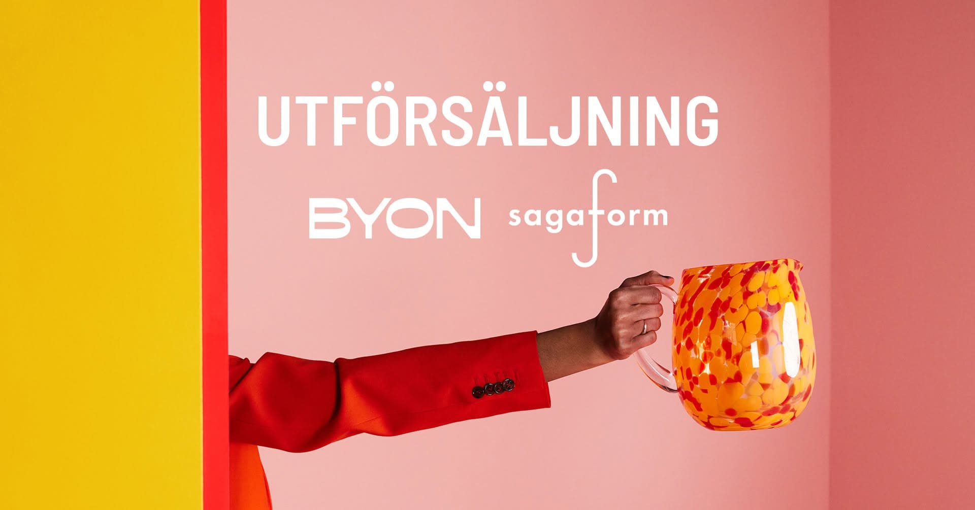 En vattenkanna i färgat glas, rött och orange, hålls upp av en utsträckt arm. Text: utförsäljning Byon Sagaform.